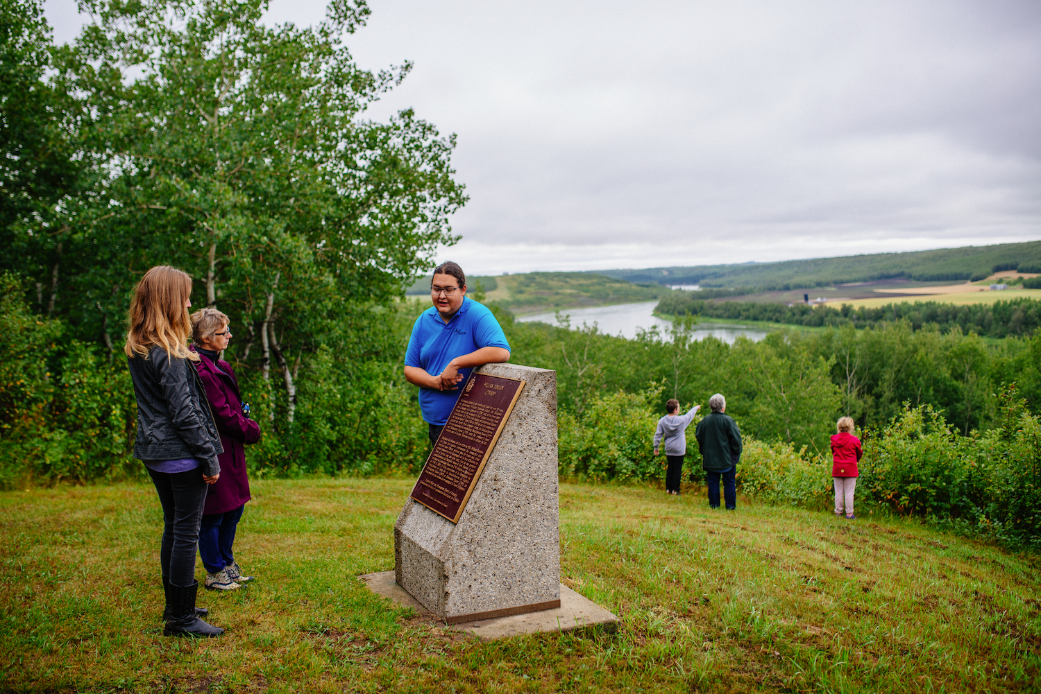 Interpreter leans on historic plaque while talking to 2 adult visitors. 2 children are exploring nearby plants in the background.