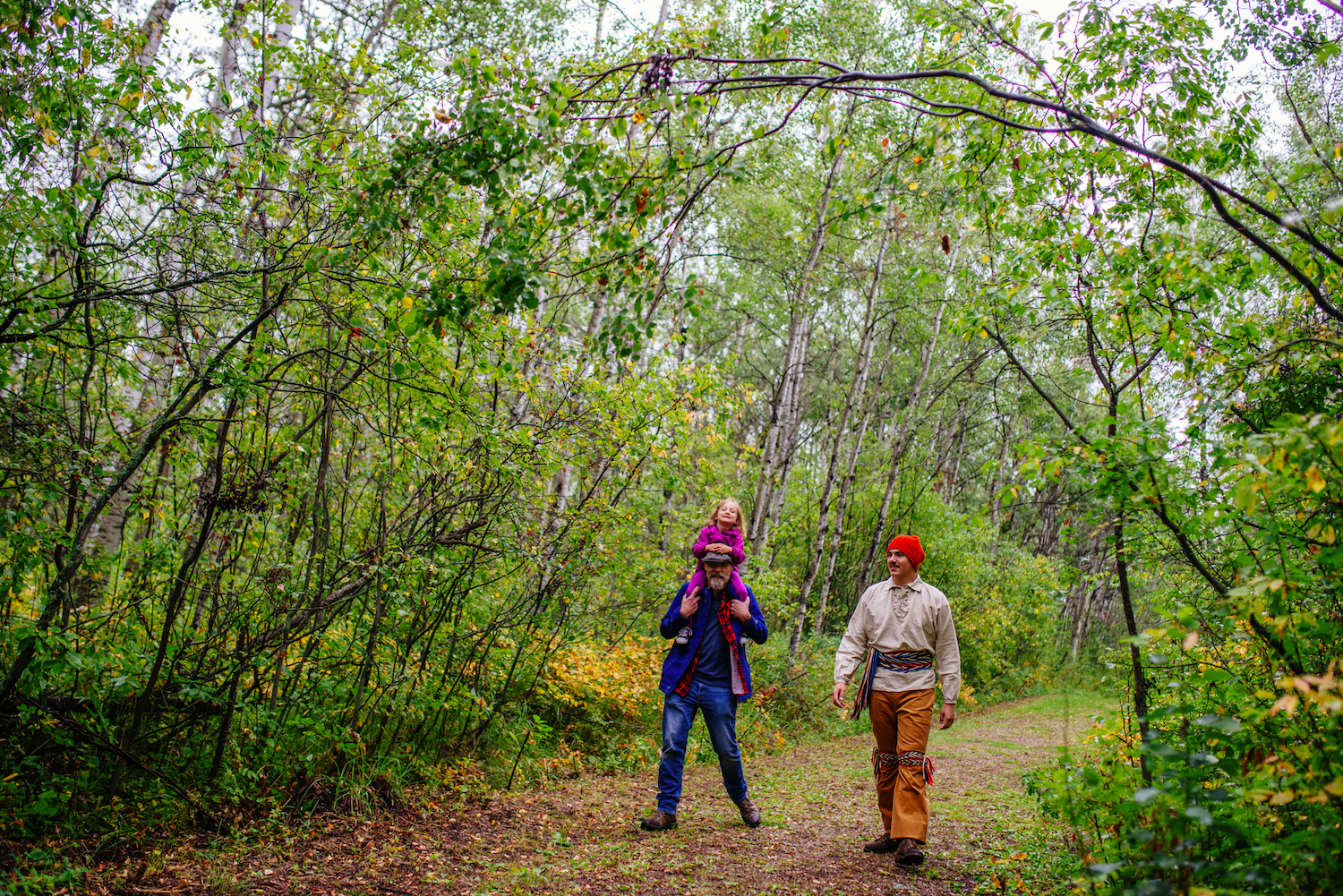 a senior visitor with a small child on his shoulders walks alongside an interpreter through a wooded trail.