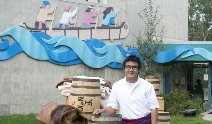 A tour guide stands outside the Fort George & Buckingham House visitor centre with a collection of fur trade items. These items include barrels, furs, a roll of birchbark, and woolen blankets.